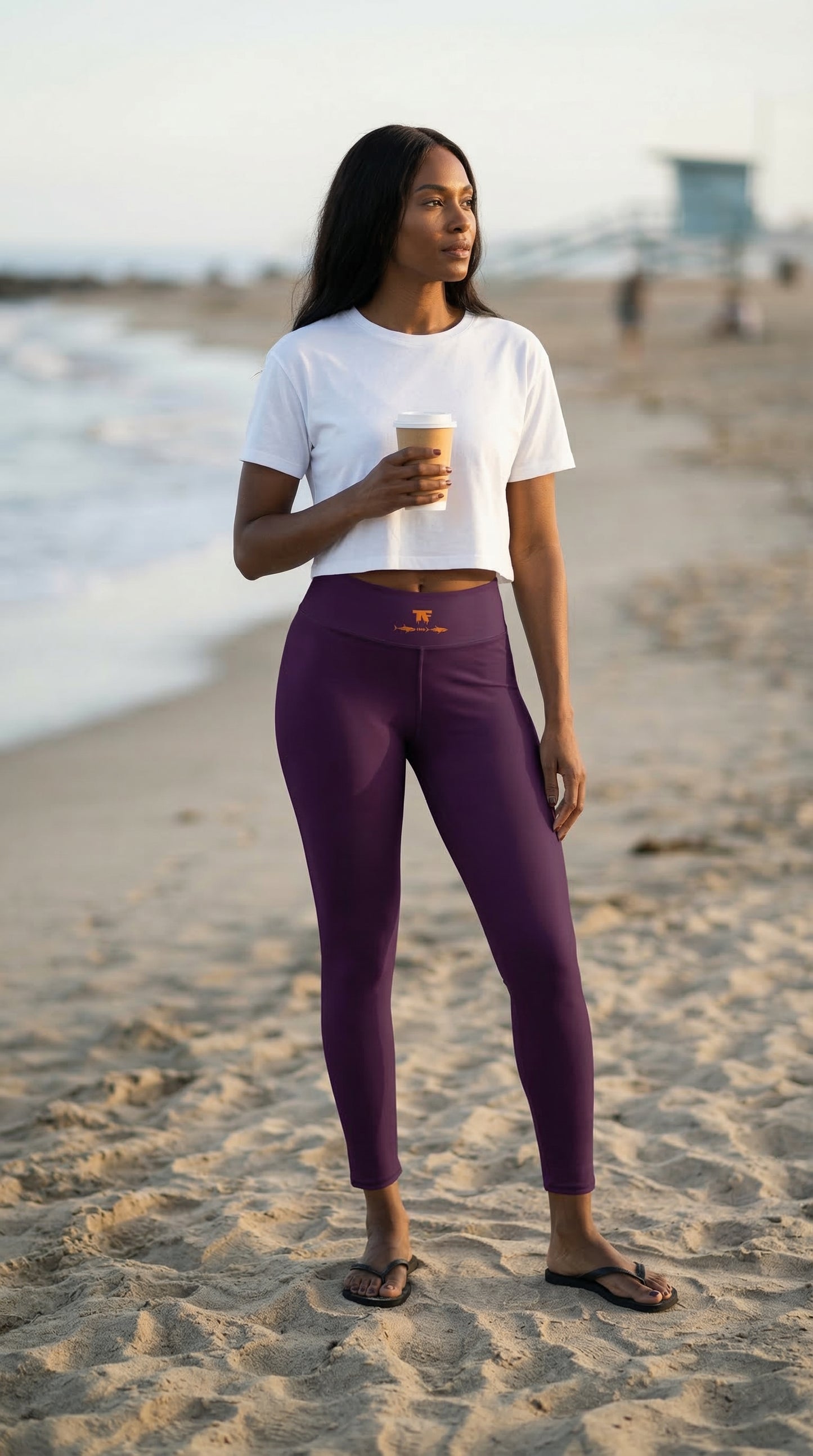 Woman in white crop top and purple leggings standing on a beach holding a coffee cup.
